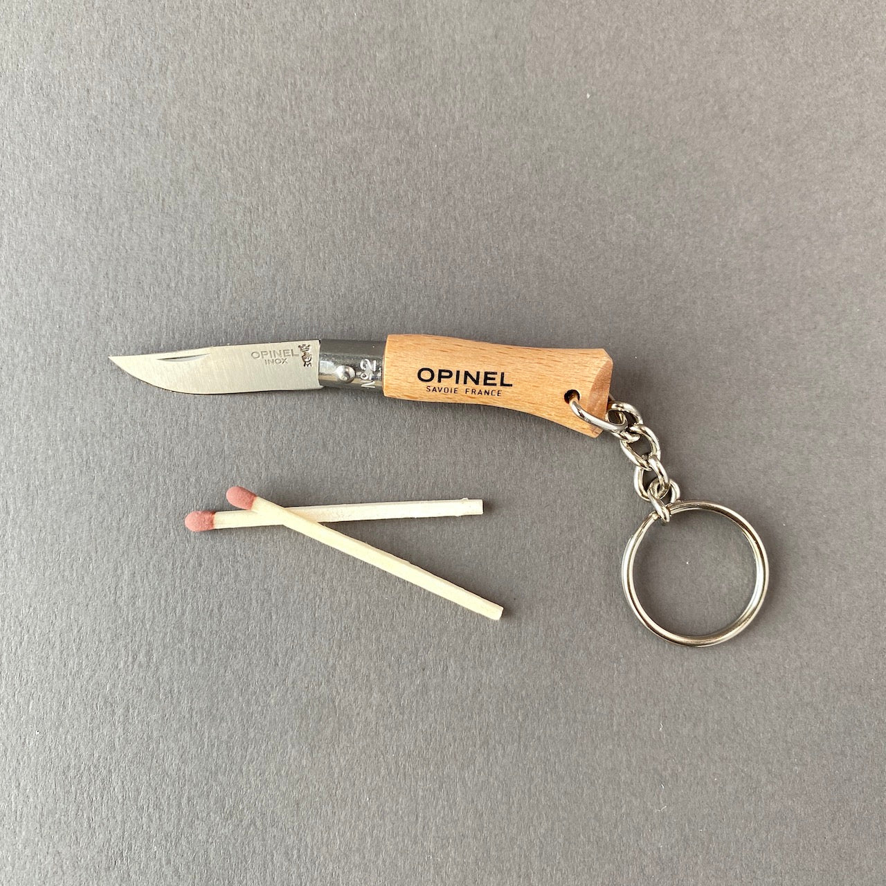 Small key chain pocket knife made out of beech wood with stainless steel blade connected to key ring with small chain-folded open with matchstick for scale reference