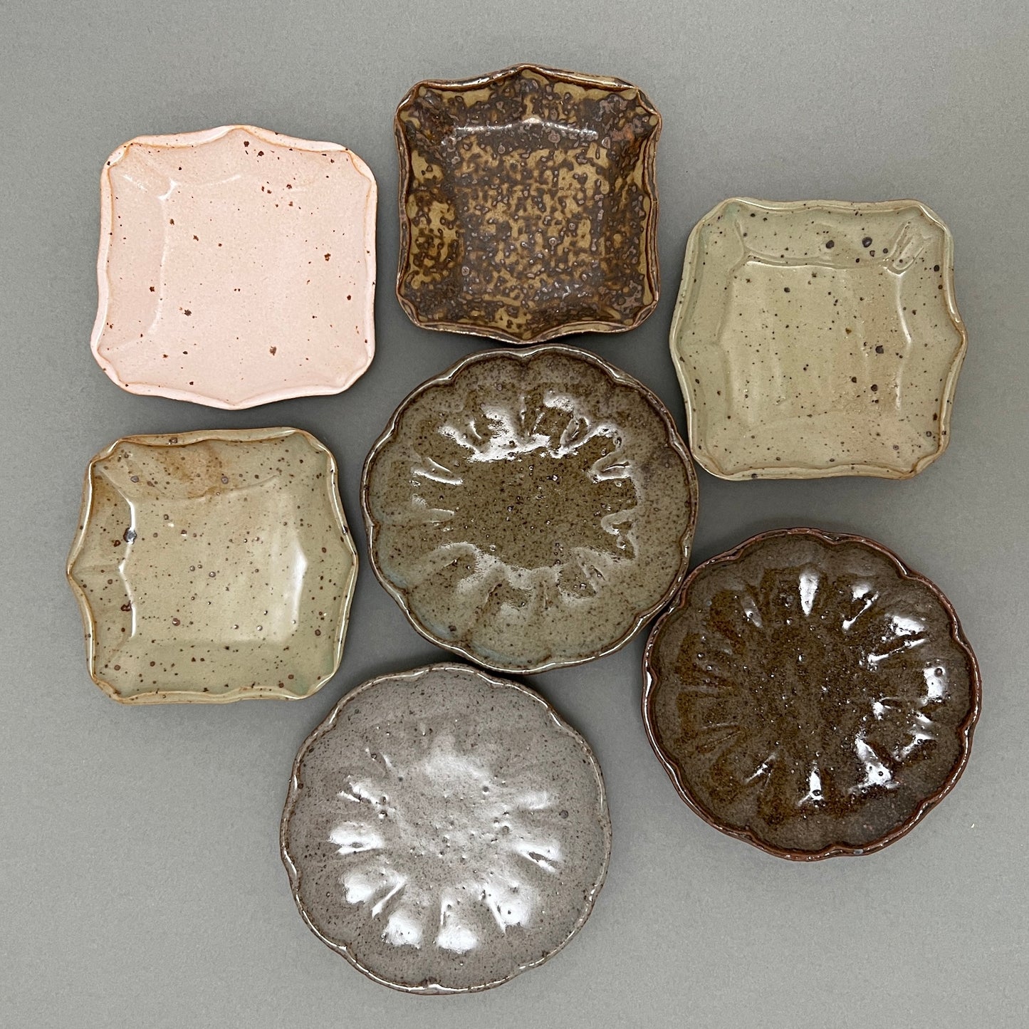 A group of seven small different color and shaped ceramic plates laying next to each other on a grey background