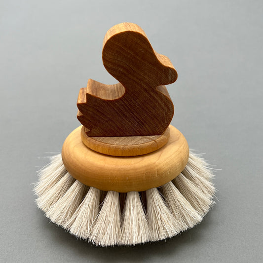 A bath brush in birch and horse hair with a wooden handle shaped like a duck. The brush is laying on a grey background