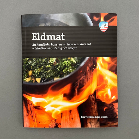 A black colored book called "Eldmat" with a picture of food cooking on a bonfire on the cover laying on a gray background