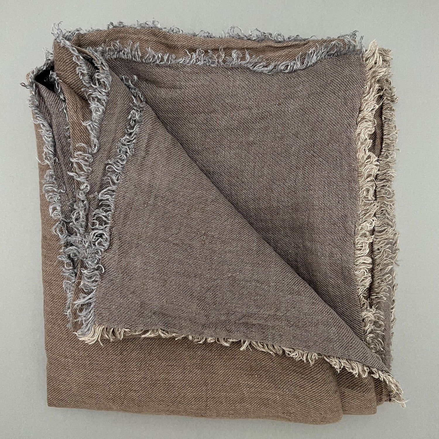 A grey/brown scarf made out of linen laying folded on a gray background