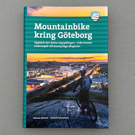  A blue colored book called "Mountainbike kring Göteborg" with a picture of a guy mountain biking in the city on the cover. Laying on a gray background