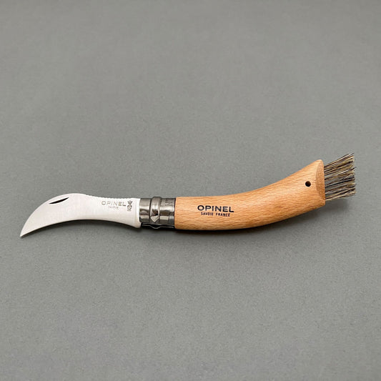 A stainless steel folding knife from Opinel with a wooden handle with a brush on the end laying on a gray background