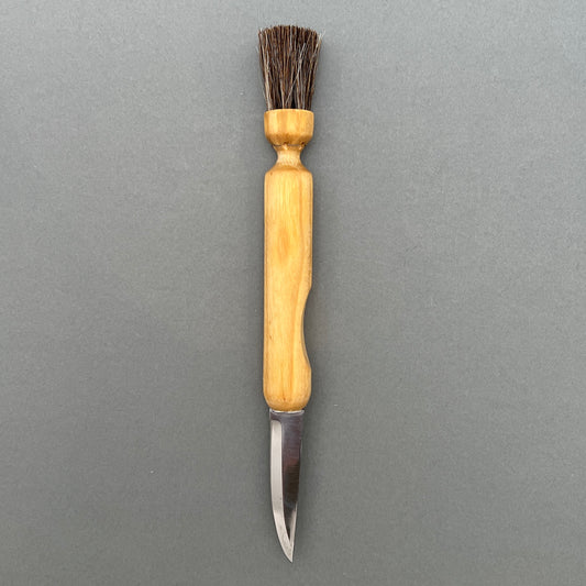 A mushroom knife and brush with handle made out of birch, a blade made out of stainless steel and a brush made out of horse hair laying on a grey background