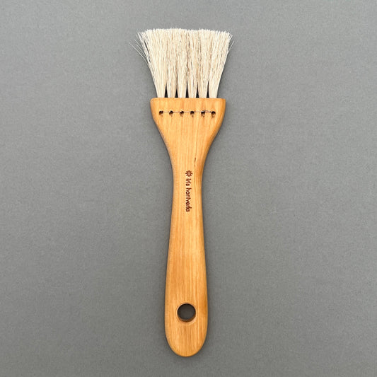 A pastry brush made out of oiled birch and horse hair with hole for hanging laying on a gray background