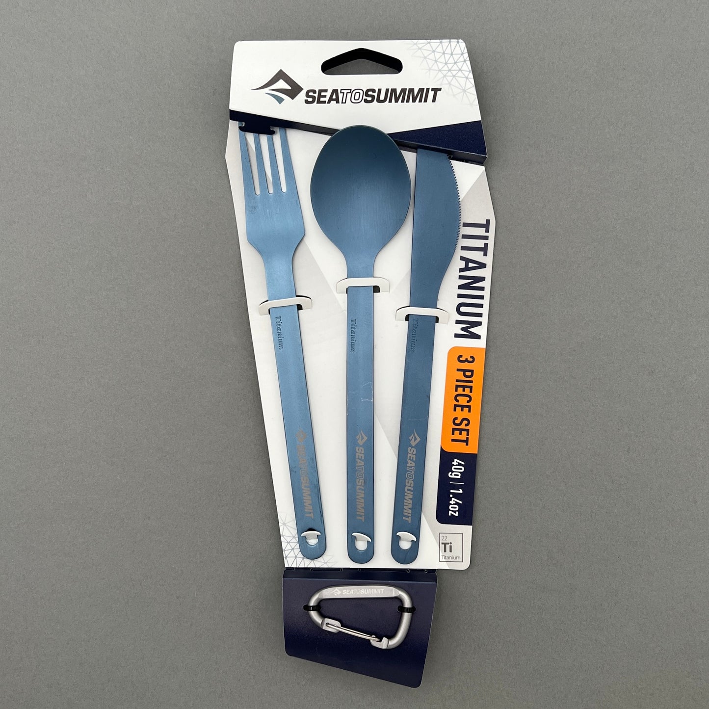 A three piece titanium cutlery set containing a fork, knife, spoon as well as a small carabiner in its package from Sea to Summit laying on a gray background