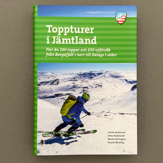 A green, blue and white book with a guy skiing downhill on the cover of a book called "Toppturer i Jämtland" laying on a gray background