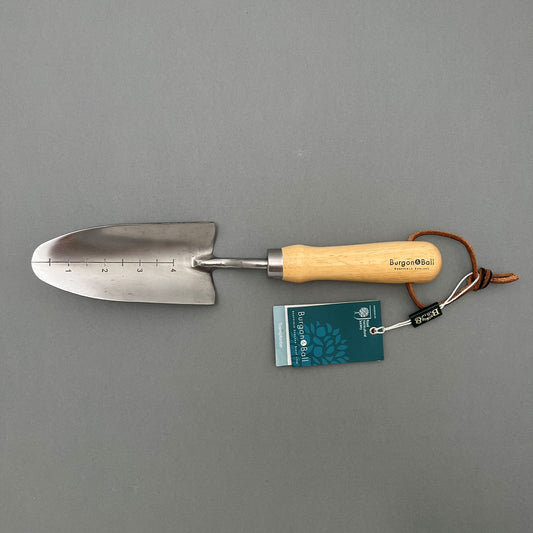 Burgon and Ball stainless steel transplanter for planting and moving plants with a wooden handle laying on a gray background