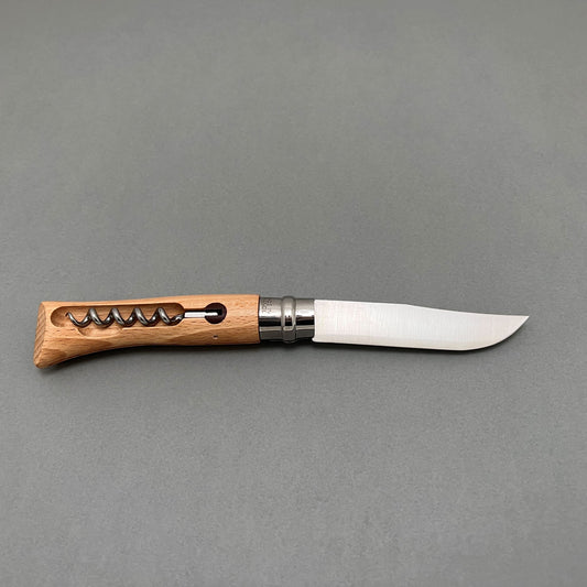 A foldable stainless steel knife from opinel with a wooden handle with an attached corkscrew laying on a gray background