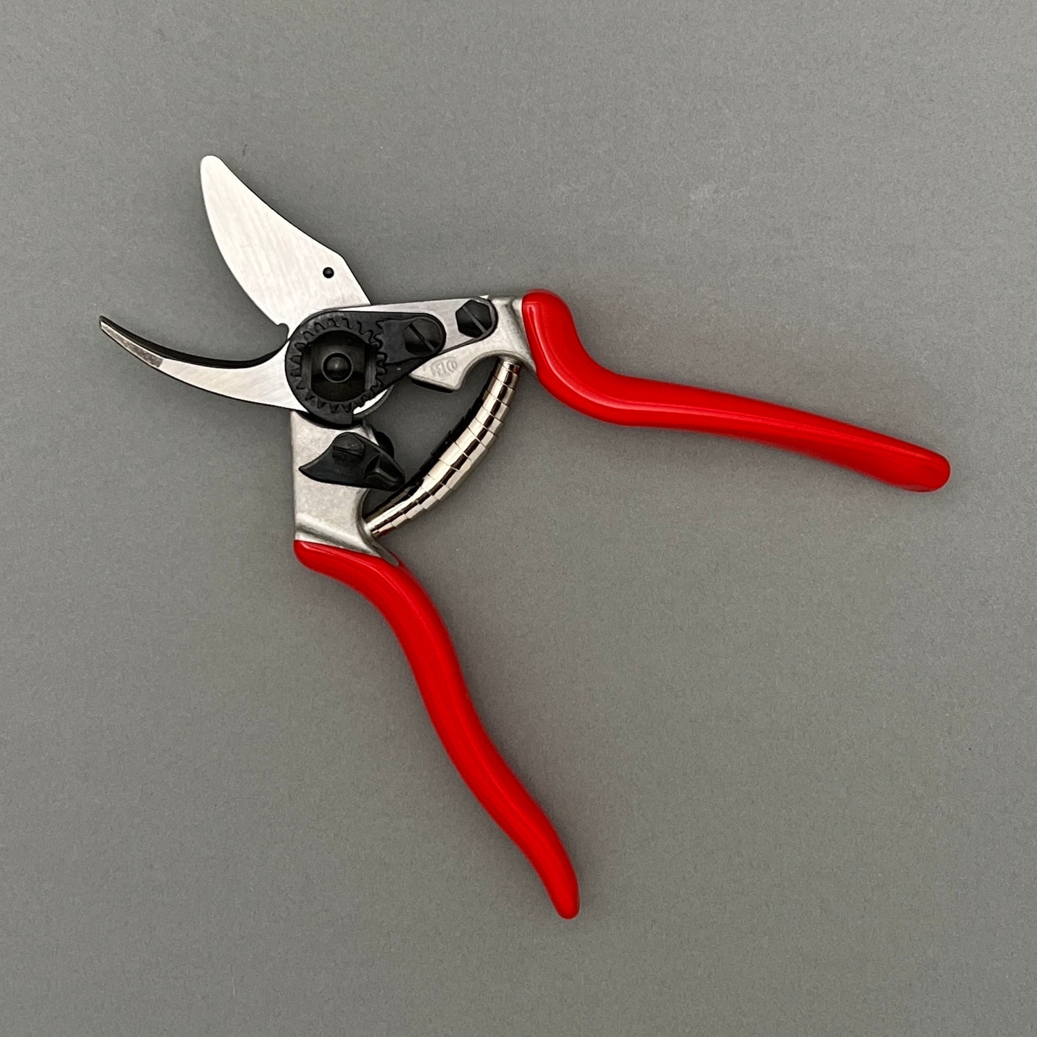 A pair of small steel clippers with a red handle in an opened position laying on a gray background