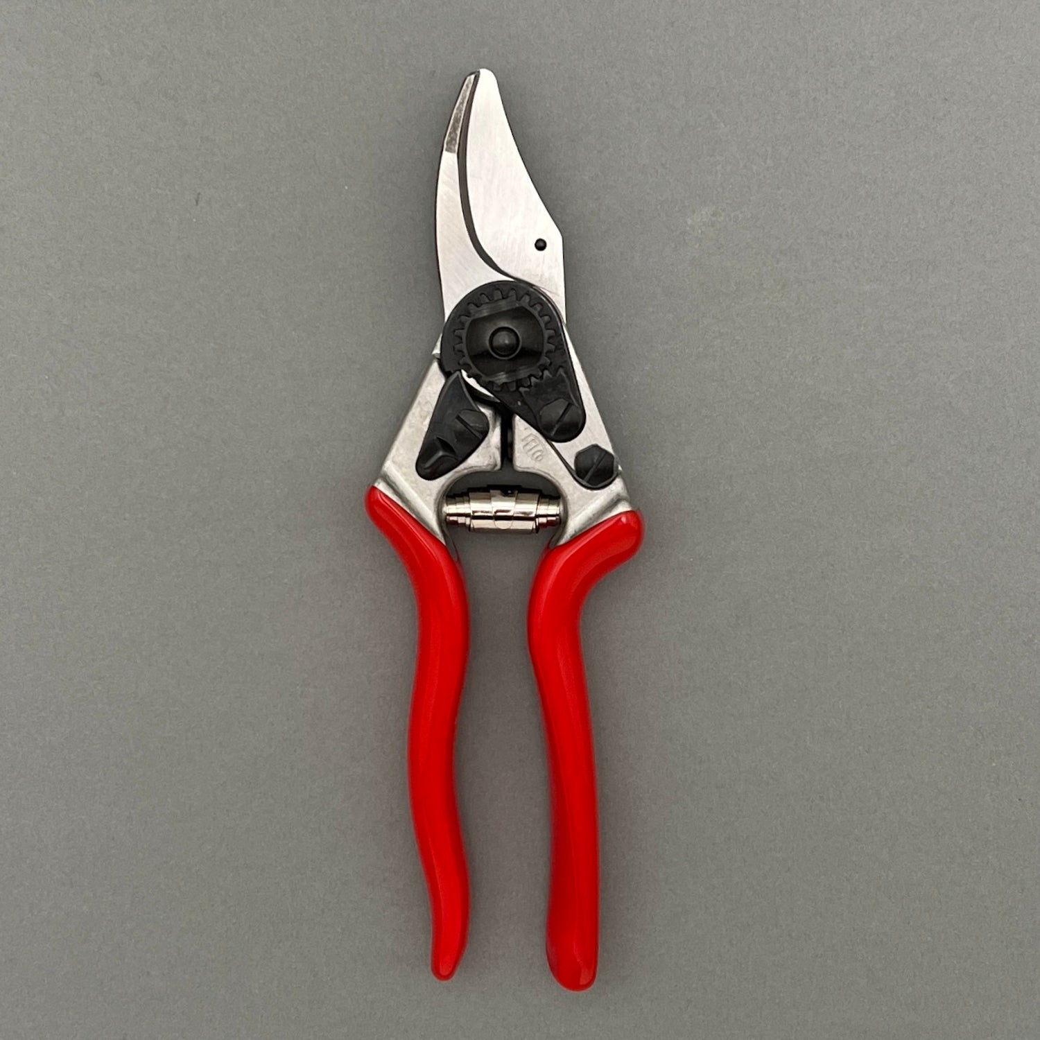 A pair of small steel clippers with a red handle in a closed position laying on a gray background